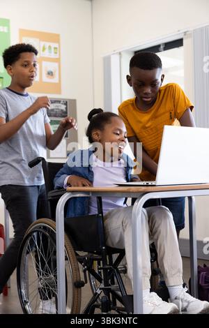 Diverse students collaborating on laptop in classroom, girl in wheelchair smiling, at school Stock Photo