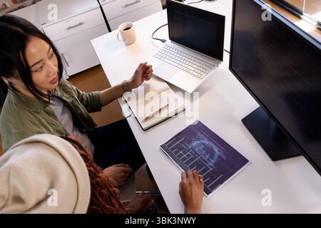 Diverse women collaborating on coding project at office desk with laptop and monitor Stock Photo