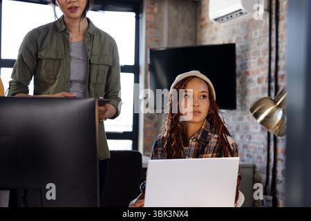 woman coding on laptop while colleague discusses project in office Stock Photo