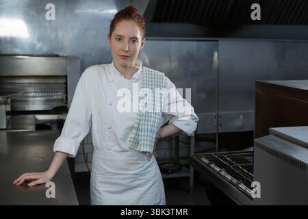 Female chef wearing chef jacket and checkered towel leaning on counter in kitchen with ovens Stock Photo