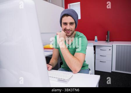Male wearing green t-shirt and beanie sitting at desk in cubicle, leaning on fist beside notebooks Stock Photo