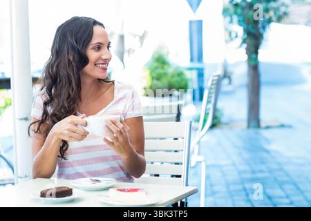 Woman sitting at metal table in sidewalk cafe holding coffee cup, smiling near pastries, copy space Stock Photo
