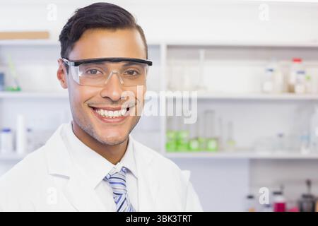 Smiling male scientist in safety goggles, lab coat, striped tie posing in lab with green solution Stock Photo