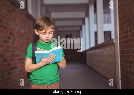 Boy student reading blue hardcover book in school corridor carrying black backpack near lockers Stock Photo