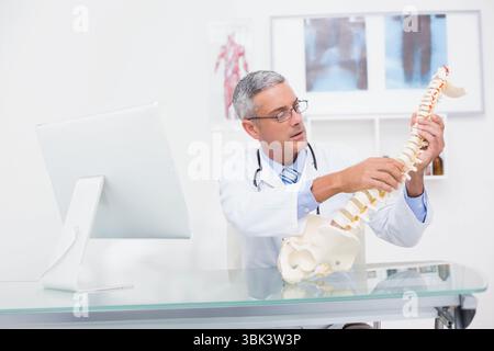 Middle-aged man doctor wearing lab coat examining spinal column model at glass-top desk in clinic Stock Photo