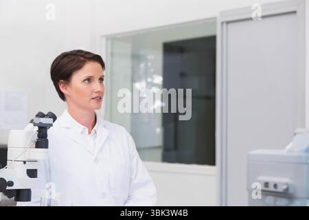 Female scientist wearing lab coat operating microscope in clinical lab beside diagnostic analyzer Stock Photo