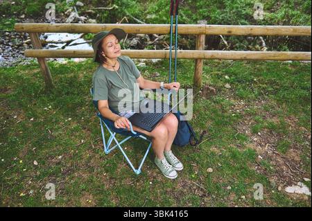 A woman enjoys a peaceful moment in nature seated on a portable chair, working on her laptop during a hiking adventure. Stock Photo