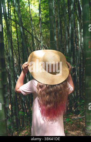 Back view of a girl with long wavy hair and a straw hat standing in a dense bamboo forest. She is wearing a light pink dress and looking up at the towering bamboo stalks surrounding her. The sunlight filters through the green canopy, creating a peaceful and immersive atmosphere. Captures the essence of nature exploration, mindful travel, and eco tourism in Asia.  Stock Photo