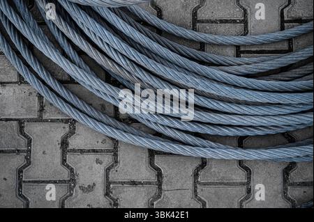 Top-down view of a thick, heavy-duty braided steel cable coiled on interlocking concrete paving stones. strength, industry, construction, durability Stock Photo
