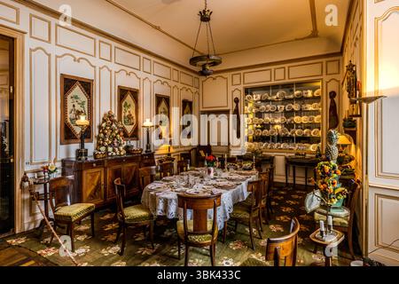 The dining room of the Château de Boussac, Creuse department, Nouvelle-Aquitaine, France, is furnished with a laid table and French porcelain. The oval table has a white lace tablecloth, plates, glasses, candlesticks and a bouquet of red flowers. Wooden chairs with green seat cushions surround it on a green carpet with a floral pattern. Rows of porcelain plates stand in an illuminated wall niche, and narrow wall hangings with figures hang on the pale wall panelling. Table centrepieces in the form of fruit and flower pyramids stand on the sideboard on the left and on a side table on the right,  Stock Photo