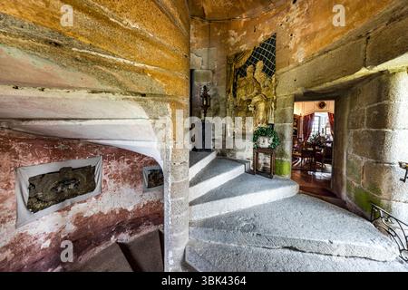 The stone spiral staircase in the side tower of the Château de Boussac, Creuse department, Nouvelle-Aquitaine, France, winds around a solid newel. Framed tapestry fragments on pale fabric hang on the red-plastered wall. At the top, a gilded relief with seated figures stands behind a grille, with a lantern on a pedestal and a plant on a small wooden table in front of it. The grey stone steps are worn. On the right, a door opens onto the dining room with a laid table and red curtains. Even the walls in the stairwell are adorned with tapestries. The castle is a medieval fortress with a rich histo Stock Photo