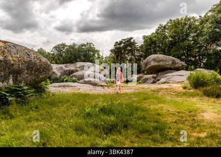 The granite blocks of Les Pierres Jaumâtres, a natural monument, lie on Mont Barlot (591 metres) near Toulx-Sainte-Croix, Creuse department, Nouvelle-Aquitaine, France, beyond a meadow. Journalist Angela Berg, wearing a straw hat, a coral-coloured wrap, a blue top and white shorts, walks across the meadow in front of the rocks. Large, rounded boulders stand on the left and in the centre, and on the right one block rests on top of flatter rocks. Pine and deciduous trees rise behind them under a cloudy sky, and ferns grow in the foreground. It is a collection of around 40 massive granite rocks f Stock Photo