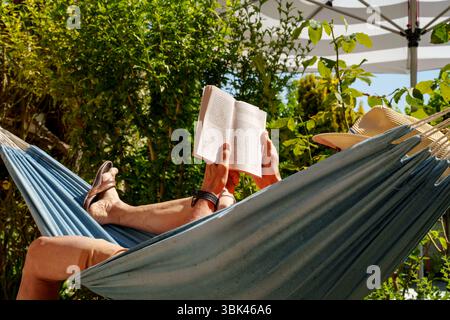 A Man Wearing A Sun Hat Reads A Book In A Hammock. Lush Garden Landscape And Striped Parasol. Vacation Atmosphere. Stock Photo