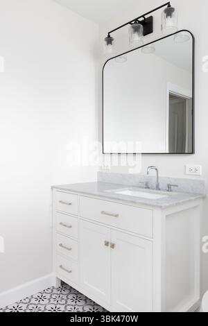 A bathroom detail with a white cabinet, grey quartz countertop, black light fixture, and a grey, white, and black pattern tile flooring. Stock Photo