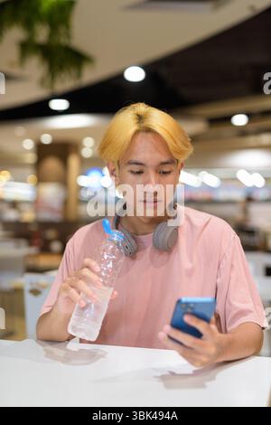 Young student teenager man with headphones drinking water and using mobile phone indoors Stock Photo