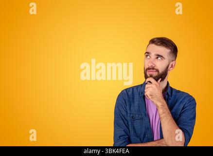 Dreamy hipster guy thinking over something, looking at empty space on orange studio background Stock Photo