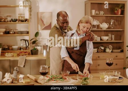 Senior Multiethnic Couple Preparing Homemade Pizza Together in Kitchen Stock Photo