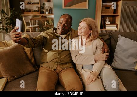 Senior Multiethnic Couple Sitting Together Taking Selfie Embracing on Sofa Stock Photo