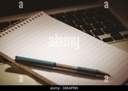 Flat design notebook with ballpoint pen is resting on laptop keyboard over desk with warm lighting Stock Photo