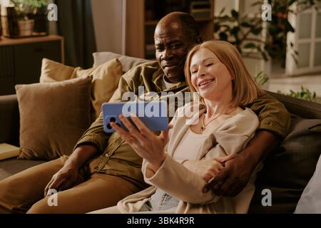 Senior Multiethnic Couple Sitting Together Smiling While Taking Selfie Indoors Stock Photo