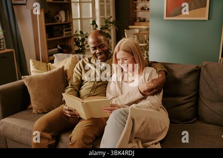 Senior Multiethnic Couple Sitting Together Reading Book and Smiling Indoors Stock Photo