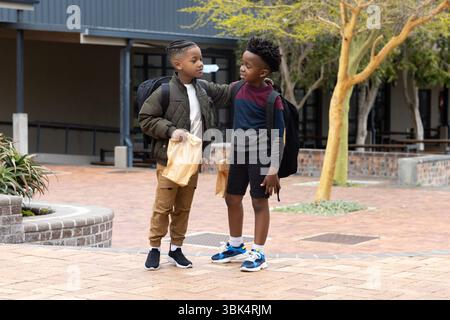 Two diverse boys with backpacks talking outside school, sharing friendly moment Stock Photo