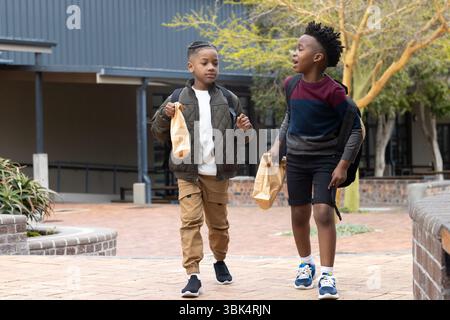 Two diverse boys walking outside school, carrying lunch bags and wearing backpacks Stock Photo