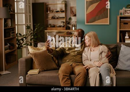 Senior Multiethnic Couple Sitting on Sofa Smiling and Taking Selfie Together Stock Photo