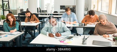 Diverse group of students sitting at desk writing in classroom Stock Photo