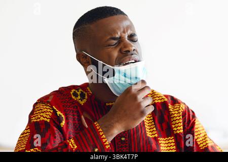 Young African Man Taking Off Medical Mask At Work, Can Not Breathe Stock Photo