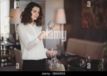 Female server wearing white blouse examining wine glasses under table lamp light in restaurant Stock Photo