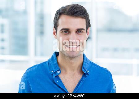 Man smiling in blue button-down shirt standing in office with large windows showing urban skyline Stock Photo