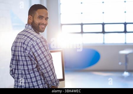 African American man standing at desk in loft office, looking over shoulder with computer monitor Stock Photo