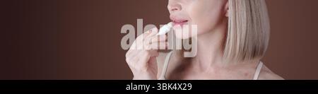 Middle-aged woman with blonde hair using lip balm applicator on plump lips. Background is blurred, bringing focus to facial features and lip balm application Stock Photo