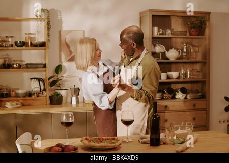 Senior Caucasian Woman and Senior Black Man Dancing Together in Kitchen Stock Photo