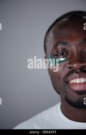 Portrait of smiling black man with green face mask applied under his left eye, focusing on skincare and self-care in close and relaxed setting Stock Photo