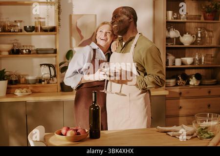 Senior Caucasian Woman and Senior Black Man Embracing and Smiling in Kitchen Stock Photo