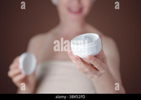 Elderly lady smiling while holding open jar of facial cream in their hand with beige background. Focus on cream jar and persons hand, face blurred in background Stock Photo
