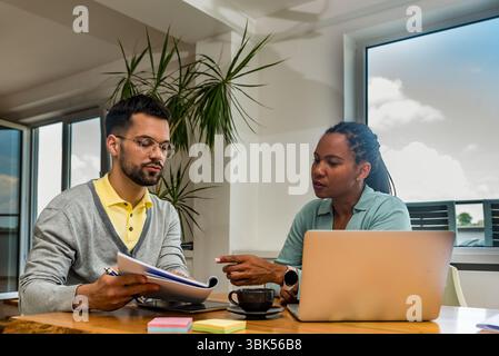 Two business professionals working together in an office reviewing documents and analyzing ideas using a laptop during a collaborative meeting focused Stock Photo