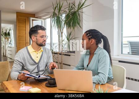Two business professionals working together in an office reviewing documents and analyzing ideas using a laptop during a collaborative meeting focused Stock Photo