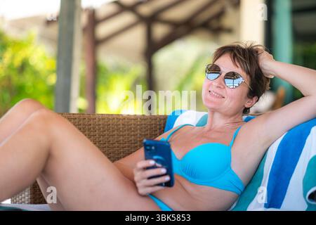Middle-aged woman in blue bikini lounging on striped towel, holding smartphone and relaxing by the pool, sunny resort atmosphere, summer holiday Stock Photo