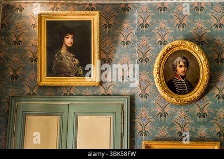 Portraits of George Sand's ancestors and descendants hang in the salon of the George Sand House (Maison de George Sand) on Place Sainte-Anne in Nohant, municipality of Nohant-Vic, in the Berry, Indre department, Centre-Val de Loire, France. On the left, a painting in a gilded frame shows a young woman with dark hair, flowers and a patterned shawl. On the right hangs an oval portrait of a man with grey hair in a dark braided uniform. A grey-green double door stands below, and the blue wallpaper has a floral pattern. The evening continued there with games and music. Some of the furniture in the  Stock Photo