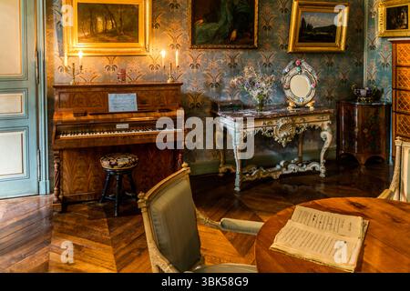 The salon in the George Sand House (Maison de George Sand) on Place Sainte-Anne in Nohant, municipality of Nohant-Vic, in the Berry, Indre department, Centre-Val de Loire, France, shows a Pleyel piano on the left with lit candles and a sheet of music, and an upholstered piano stool in front of it. A marble console table with curved legs, a bouquet of flowers and a mirror in a floral frame stands in the centre. A chest of drawers with inlay work stands on the right. In the foreground, open sheets lie on a table of reddish wood, with an armchair beside it. Paintings in gilded frames hang on the  Stock Photo