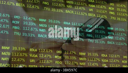 Showing hand holding smartphone above laptop keys with mug on desk, featuring stock ticker overlay Stock Photo