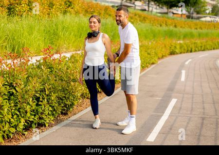 In a lively park, a fitness trainer assists a woman in stretching to enhance flexibility and promote an active lifestyle amidst lush greenery. Stock Photo