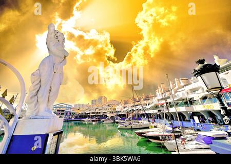 Malaga Benalmadena Marina showing white buildings with yachts  and bars and a female statue at the entrance against a golden   cloudy sky with sunbeam Stock Photo