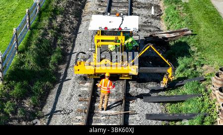 Two workers are engaged in repairing railway tracks using specialized heavy machinery in a sunny rural setting. The grass and wooden beams surround the tracks, indicating ongoing maintenance efforts. Stock Photo