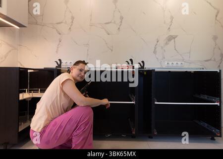 Smiling woman with screwdriver assembling kitchen cabinet, confident DIY process, modern interior with marble wall, drawer runners and carousel shelf Stock Photo