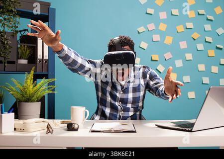 Amazed businessman using VR headset at his desk, fully engaged in a gaming session. Male entrepreneur with his mouth opened, wearing virtual reality goggles for an immersive simulation experience. Stock Photo