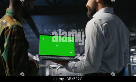 Army tech specialist in data center and coworkers using green screen laptop to troubleshoot military computer systems. Soldier and command center server room admins using chroma key notebook Stock Photo